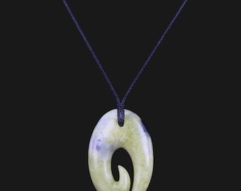 Puede incluir: Un colgante de jade verde y blanco con forma de gancho, colgado de un cordón negro.