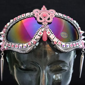 Puede incluir: Gafas de protecci&oacute;n con tachuelas rosas y plateadas, lentes iridiscentes y una corona de piedras rosas. Las gafas tienen pendientes colgantes rosas y plateados.