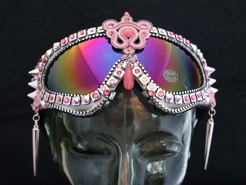 Puede incluir: Gafas de protecci&oacute;n con tachuelas rosas y plateadas, lentes iridiscentes y una corona de piedras rosas. Las gafas tienen pendientes colgantes rosas y plateados.