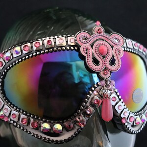 Puede incluir: Gafas de esqu&iacute; rosas y plateadas con una lente iridiscente arco&iacute;ris, adornadas con diamantes de imitaci&oacute;n y un dije de cuentas rosas.