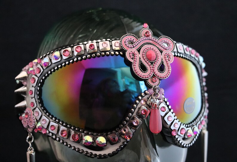 Puede incluir: Gafas de esqu&iacute; rosas y plateadas con una lente iridiscente arco&iacute;ris, adornadas con diamantes de imitaci&oacute;n y un dije de cuentas rosas.