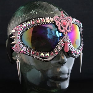 Puede incluir: Gafas de esqu&iacute; rosas y plateadas con pedrer&iacute;a y un colgante de cuentas rosas. Las gafas tienen una lente iridiscente arco&iacute;ris.