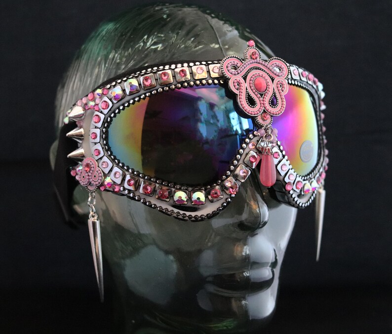 Puede incluir: Gafas de esqu&iacute; rosas y plateadas con pedrer&iacute;a y un colgante de cuentas rosas. Las gafas tienen una lente iridiscente arco&iacute;ris.