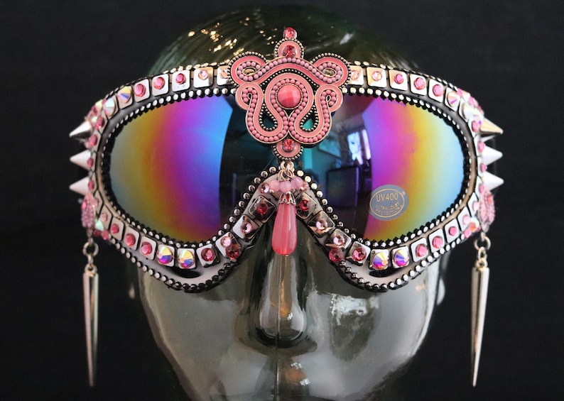 Puede incluir: Gafas de esqu&iacute; con tachuelas rosas y plateadas con un adorno de joyas rosas y pendientes colgantes. Las gafas tienen una lente iridiscente arco&iacute;ris y una etiqueta UV400.