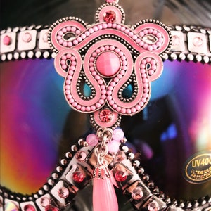 Puede incluir: Gafas de esqu&iacute; rosas y plateadas con un adorno de joyas rosas y plateadas. Las gafas tienen una lente iridiscente arco&iacute;ris y un colgante rosa y plateado.