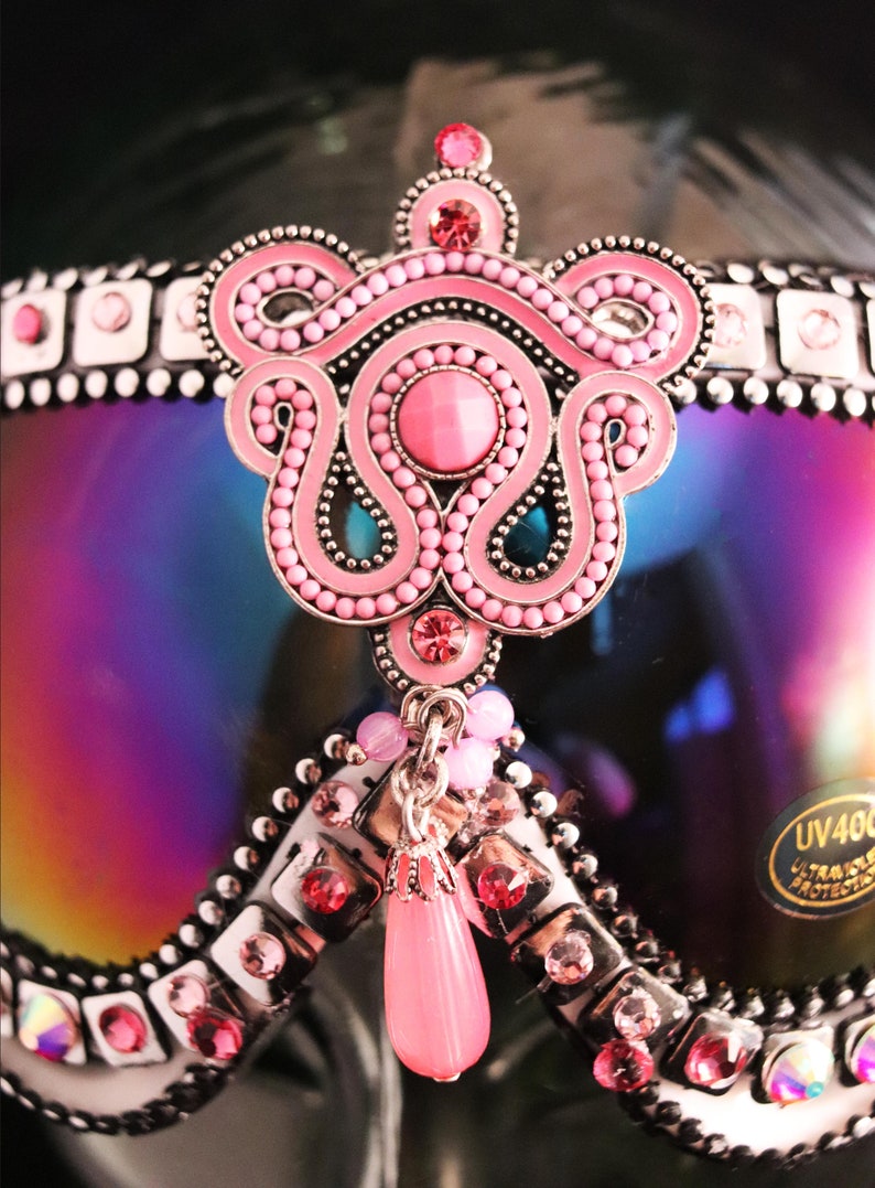 Puede incluir: Gafas de esqu&iacute; rosas y plateadas con un adorno de joyas rosas y plateadas. Las gafas tienen una lente iridiscente arco&iacute;ris y un colgante rosa y plateado.