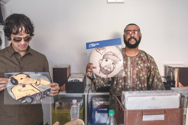 May include: Two people are holding vinyl records. The person on the left is holding a record with a cartoon illustration of a person with an elephant trunk. The person on the right is holding a record with a cartoon illustration of a person wearing a hat. The record is in a clear plastic sleeve with a blue label that says "Local Jazz".