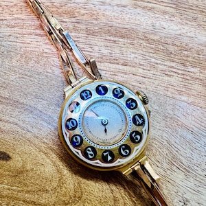 May include: A gold wristwatch with a round face and a unique design. The watch face has black Roman numerals and a white background. The watch band is made of gold metal and has a clasp closure.