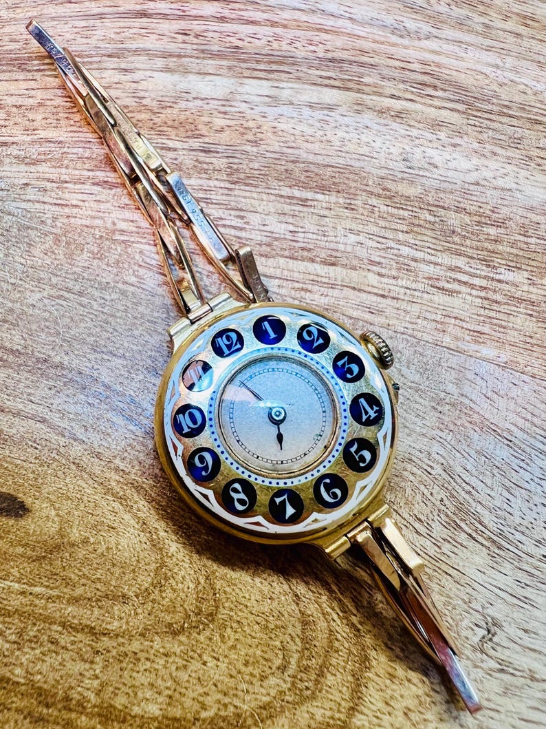 May include: A gold wristwatch with a round face and a unique design. The watch face has black Roman numerals and a white background. The watch band is made of gold metal and has a clasp closure.