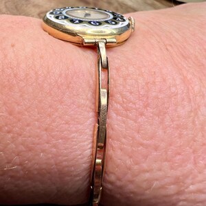 May include: A gold-toned wristwatch with a round face and black Roman numeral hour markers. The watch has a white dial with black hands and a gold-toned bracelet.