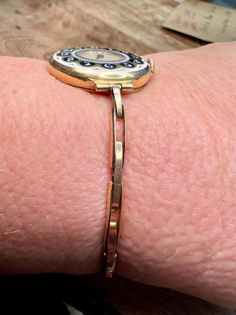 May include: A gold-toned wristwatch with a round face and black Roman numeral hour markers. The watch has a white dial with black hands and a gold-toned bracelet.