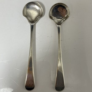 May include: A pair of silver spoons featuring a unique, scalloped bowl shape. The spoons have long, thin handles, with the number "1935" engraved near the base of each. They are set against a plain, light-coloured backdrop.