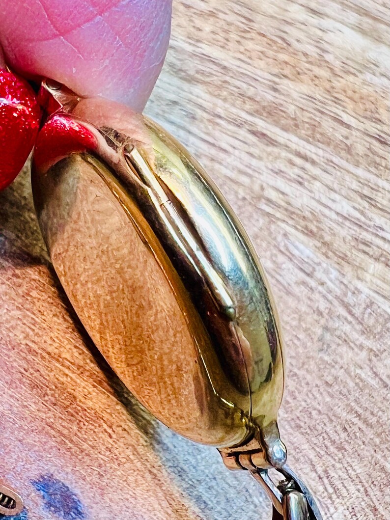 May include: A gold-toned pocket watch with a hinged lid. The watch is closed and the lid is facing the camera. The watch is on a wooden surface.