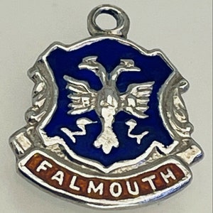 Könnte beinhalten: Silberner Anhänger mit einem blauen Emaille-Schild, auf dem ein weißer Vogel mit ausgebreiteten Flügeln abgebildet ist. Das Schild ist von einer silbernen Schriftrolle mit dem Wort "FALMOUTH" in orangefarbenem Emaille umgeben.