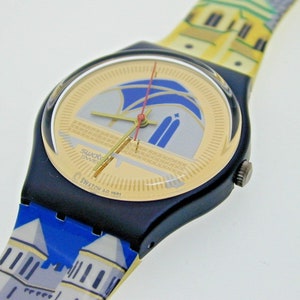Könnte beinhalten: Eine schwarze und goldene Swatch-Uhr mit einem blau-weiß gemusterten Armband. Das Zifferblatt der Uhr hat einen goldenen Hintergrund mit einem blau-weißen Design. Das Armband hat ein Muster aus Gebäuden. Das Zifferblatt der Uhr zeigt "Swatch AG 1987".