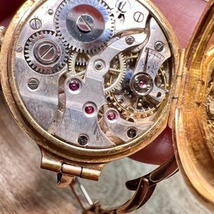 May include: A gold-toned wristwatch with a round face and a hinged back. The back is open, revealing the intricate inner workings of the watch, including gears and a small, circular dial. The watch has a gold-toned band with a clasp.