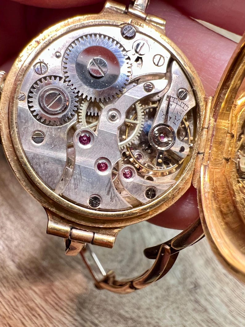 May include: A gold-toned wristwatch with a round face and a hinged back. The back is open, revealing the intricate inner workings of the watch, including gears and a small, circular dial. The watch has a gold-toned band with a clasp.