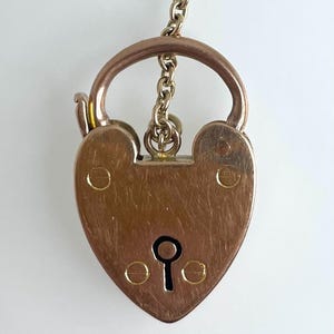 Könnte beinhalten: Ein goldfarbenes herzförmiges Vorhängeschloss mit einem Schlüsselloch und einer daran befestigten Kette. Das Vorhängeschloss hat einen abgenutzten, antiken Look.