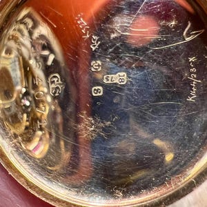 May include: Close-up of the back of a gold pocket watch with the inscription "K.W.H. 23" and the numbers "18" and "8" visible.