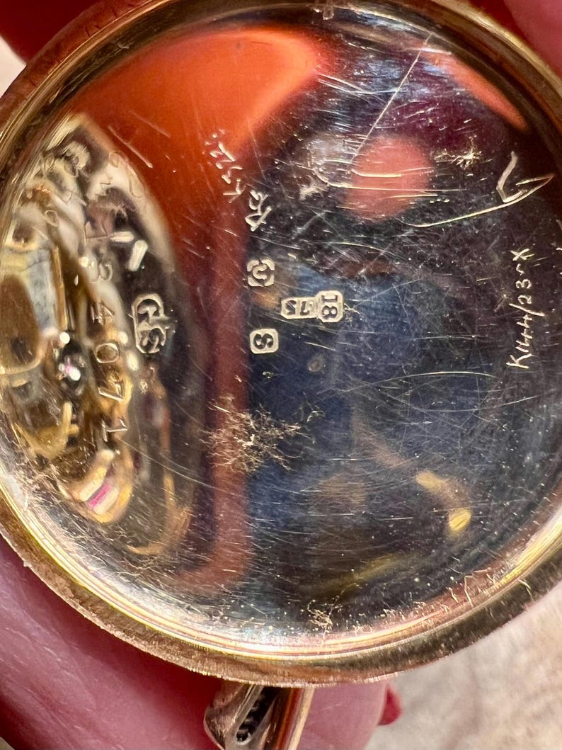May include: Close-up of the back of a gold pocket watch with the inscription "K.W.H. 23" and the numbers "18" and "8" visible.