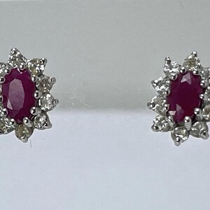 Peut inclure: Une paire de boucles d'oreilles à clous avec des pierres précieuses ovales rouge foncé, chacune entourée de petites pierres précieuses claires. Les boucles d'oreilles sont serties dans un métal argenté, créant un design floral étincelant. Bijoux classiques.