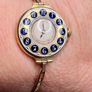 May include: A gold-toned wristwatch with a white face and blue and white enamel numerals. The watch has a bangle-style band.