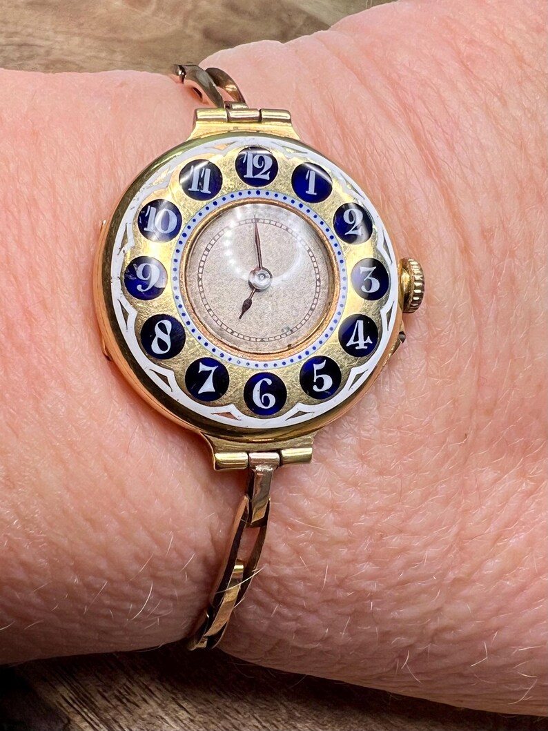 May include: A gold-toned wristwatch with a white face and blue and white enamel numerals. The watch has a bangle-style band.