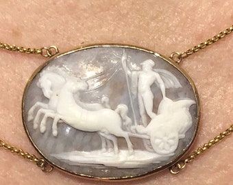 May include: A gold chain necklace with a cameo pendant featuring a white cameo carving of a figure riding in a chariot pulled by two horses.
