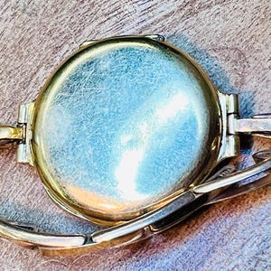 May include: A gold-toned wristwatch with a round face and a metal band. The watch face is smooth and has a slight sheen.