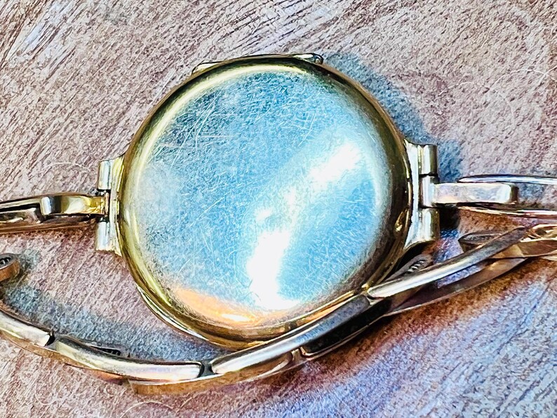 May include: A gold-toned wristwatch with a round face and a metal band. The watch face is smooth and has a slight sheen.