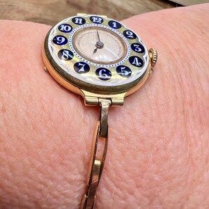 May include: A gold wristwatch with a white face and blue and white numbers. The watch has a gold band.