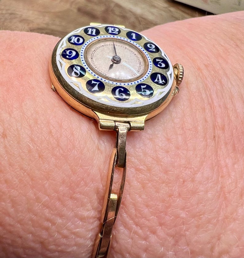 May include: A gold wristwatch with a white face and blue and white numbers. The watch has a gold band.