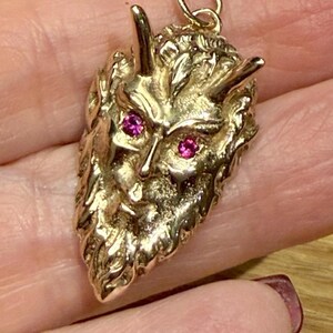 May include: A gold-coloured pendant shaped like a horned demon face, featuring two red gemstones as eyes. The pendant has a textured surface and a small loop at the top for a chain.