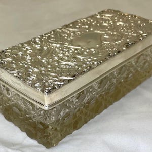 May include: A rectangular silver box with an ornate, embossed lid. The lid has floral patterns and an oval centre. The box's body is clear, textured glass, giving it a vintage appearance. The box is approximately 15 cm long.
