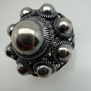 Op de afbeelding: Een zilverkleurige metalen ring met een cluster van bolvormige en afgeronde elementen. De ring heeft een grote centrale bol omringd door kleinere bollen, allemaal verbonden door ingewikkeld, getextureerd metaalwerk. Het ontwerp heeft een vintage uitstraling.