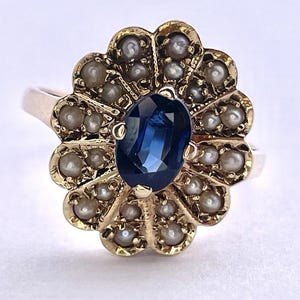 Könnte beinhalten: Ein goldfarbener Ring mit einem großen, ovalen, dunkelblauen Edelstein in der Mitte, umgeben von einem floralen Arrangement aus kleinen Perlen. Das Design des Rings ist aufwendig und detailliert, mit Vintage-Ästhetik.