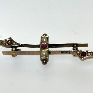 May include: A gold-toned brooch featuring a central cross design. The cross is decorated with small white pearls and red gemstones. The brooch has two curved arms extending out from the cross.