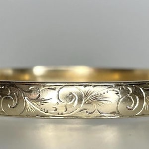 Peut inclure: Un bracelet jonc doré avec un motif floral gravé. Le bracelet est doté d'une fermeture à fermoir et d'une finition lisse et polie. Le design complexe présente des feuilles tourbillonnantes et des détails délicats.