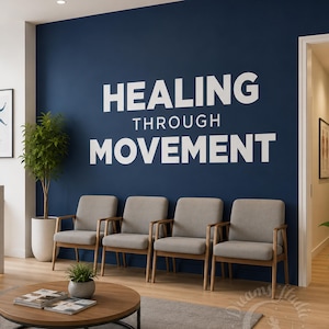 May include: A waiting room featuring a dark blue wall with the white text HEALING THROUGH MOVEMENT. Four light gray chairs with wooden frames are arranged in a row. A round wooden coffee table sits in the foreground with two books.