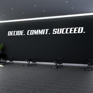 以下が含まれることがあります： 黒い壁に、太字のサンセリフ体で白い文字「DECIDE. COMMIT. SUCCEED.」が書かれています。壁の下にはダークグレーの床があり、黒いエクササイズベンチが並んで配置されています。左側には小さな緑の植物が見えます。