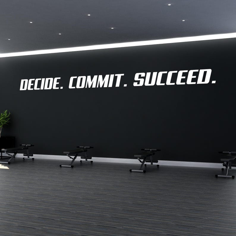 以下が含まれることがあります： 黒い壁に、太字のサンセリフ体で白い文字「DECIDE. COMMIT. SUCCEED.」が書かれています。壁の下にはダークグレーの床があり、黒いエクササイズベンチが並んで配置されています。左側には小さな緑の植物が見えます。