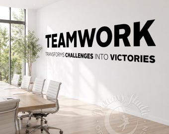 Puede incluir: Un adhesivo de pared negro con la palabra TEAMWORK en letras mayúsculas grandes, seguido del texto TRANSFORMS CHALLENGES INTO VICTORIES en una fuente más pequeña. Las letras están aplicadas sobre una pared blanca en una oficina.