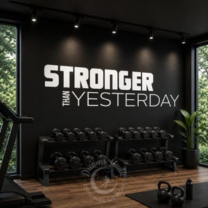 Könnte beinhalten: Ein schwarzer Wandaufkleber mit dem weißen Text STRONGER THAN YESTERDAY in einer fetten serifenlosen Schrift. Der Aufkleber ist an einer dunklen Wand über zwei Metallregalen angebracht, die Hanteln in einem Fitnessraum beherbergen.