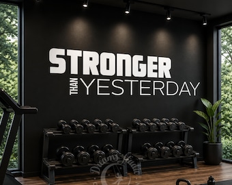 以下が含まれることがあります： 太字のサンセリフ体で「STRONGER THAN YESTERDAY」と白文字で書かれた黒いウォールステッカー。このステッカーは、複数の黒いダンベルが置かれた2つの金属製ダンベルラックの上の暗い壁に貼られています。