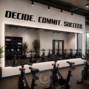 Könnte beinhalten: Ein großer Wandaufkleber mit schwarzen Großbuchstaben, der DECIDE. COMMIT. SUCCEED. lautet und an einer weißen Wand über einem breiten rechteckigen Spiegel in einem Fitnessstudio angebracht ist. Der Spiegel reflektiert mehrere Heimtrainer und Pflanzen.