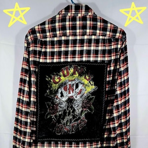 Pode incluir: Uma camisa de flanela de manga comprida com botões e um padrão xadrez preto, vermelho e branco. A parte de trás apresenta um patch preto com um gráfico Guns N' Roses, incluindo texto e imagens de armas e rosas.