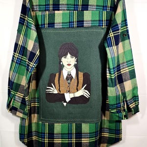 May include: Green, blue, and tan plaid flannel shirt with a dark green patch featuring a cartoon illustration of a person with dark braids, wearing a black dress and brown vest. The shirt has long sleeves.