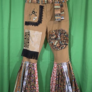 May include: Tan pants featuring a decorative belt, a black lace patch with a bee embroidery, a fabric ruffle, and a black fringe accent. The flared leg openings are embellished with multiple panels of patterned fabric in floral and paisley designs.