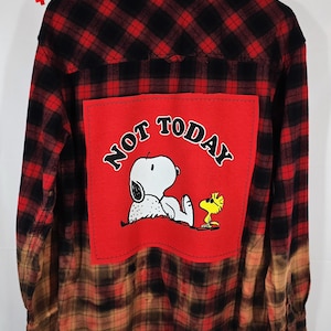 以下が含まれることがあります： 赤と黒のチェック柄フランネルシャツ、下半分は漂白されています。「NOT TODAY」の文字とスヌーピーとウッドストックが描かれた赤い正方形のパッチ。2つのピクセル化された赤いハートが上隅にあります。
