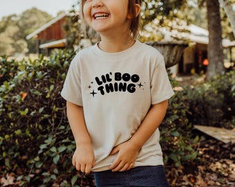 Può includere: Una t-shirt a maniche corte color beige chiaro con il testo nero LIL' BOO THING stampato in un carattere grassetto e arrotondato sul petto, accentuato da quattro piccoli simboli a stella a quattro punte che circondano la scritta.